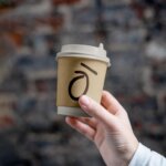 Hand holding Kohi branded takeaway coffee cup against textured wall background